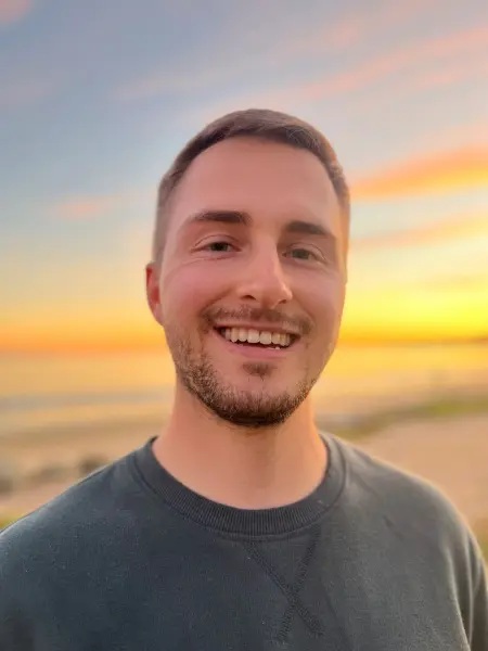 Close up photograph of Kyle Mayl smiling and standing in front of a sunset.